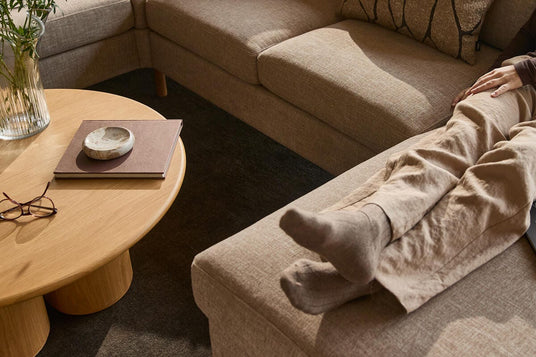 Beige hörnsoffa med kuddar; person med strumpor vilar benen på schäslongen. Rundt soffbord i trä med vas, glasögon och bok i ett varmt, neutralt vardagsrum.
