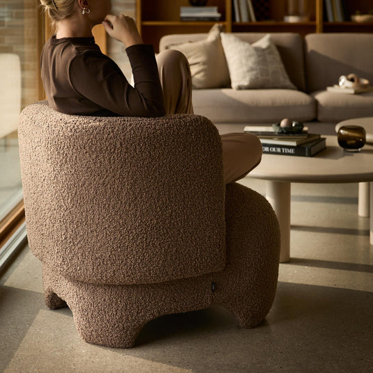En person sitter i en brun bouclé‑fåtölj i ett mysigt vardagsrum med beige soffa och runt soffbord med böcker, varav en har titeln ”OUR TIME”.