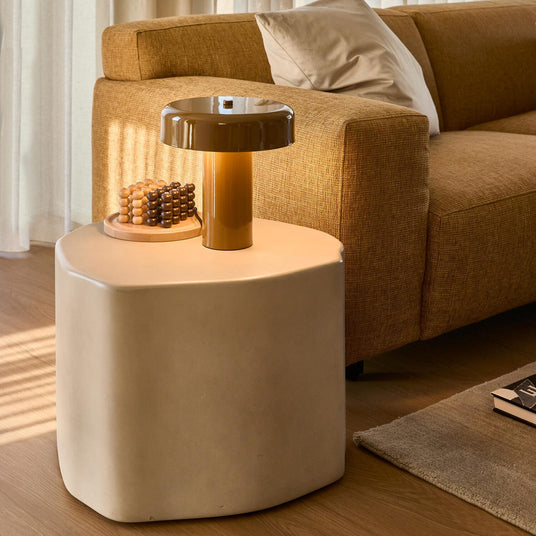 Vardagsrum i varma toner med skulpterat beige sidobord, tänd karamellfärgad svamplampa, träpärlor på bricka och senapsgul soffa med kudde vid skira gardiner i dagsljus.