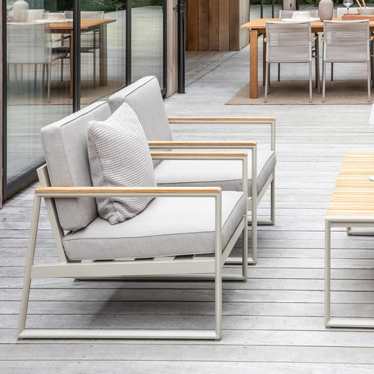 Modern uteplats med ljusgrå loungesoffor, soffbord i trä och glasfasad med utsikt mot grönskande träd.