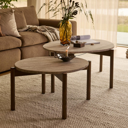 Två runda soffbord i trä på en ljus strukturmatta, med bärnstensfärgad vas med grenar och ett tänd ljus; beige soffa och skira gardiner i dagsljus.