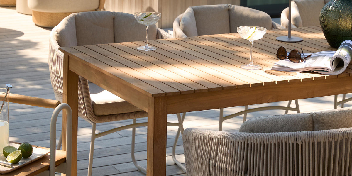 Solig terrass med matbord i trä och beige repstolar; två drinkglas, liten bordslampa, grön vas och uppslagen tidning på bordet.