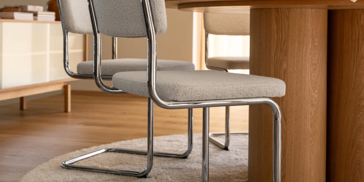 Två ljusgrå stoppade cantilever-stolar med krom vid ett runt matbord i ek på en beige matta i en ljus matsal.