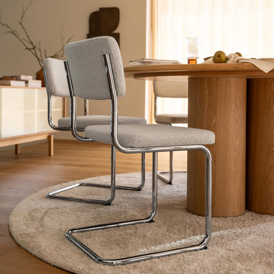 Två ljusgrå stoppade cantilever-stolar med krom vid ett runt matbord i ek på en beige matta i en ljus matsal.