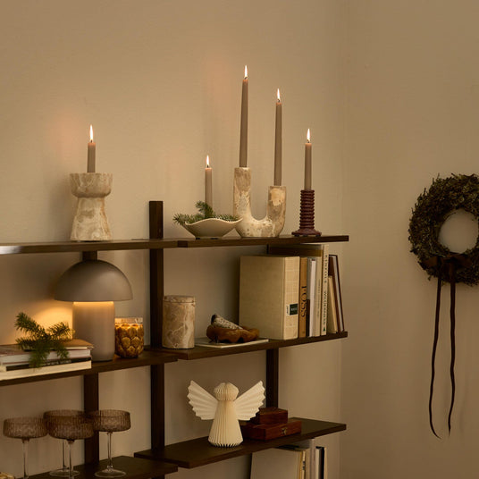Mörk trä-hyllvägg med tända ljus i ljusstakar, en bordslampa som lyser varmt, böcker och glas; en grön krans hänger på väggen.