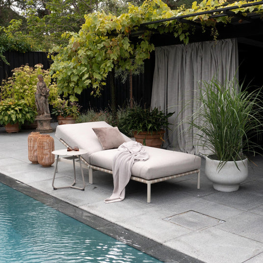 Poolsida med solstol i beige med kudde och stickad pläd, litet sidobord och rottinglyktor under en vinrankeklättrande pergola med krukväxter vid en pool.