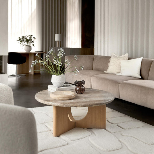 Ljus skandinavisk vardagsrumsscen med beige modulsoffa, runt soffbord i trä och sten med vas med vita blommor, liten brun glasdekoration och skål på böcker ("lifestyle living"), ljusa gardiner och texturerad vit matta.