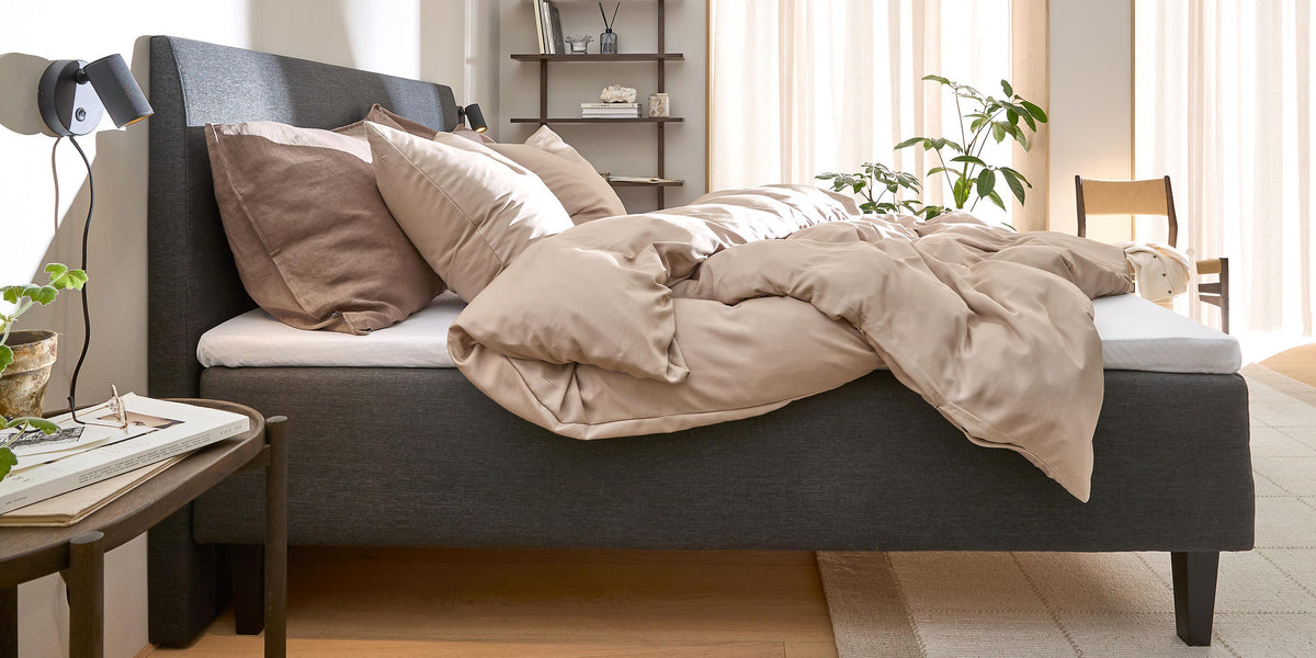 Ljust sovrum med grå stoppad säng, beige bäddset, höga tunna gardiner, vägglampor och gröna växter.