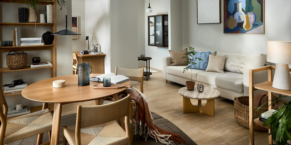 Skandinaviskt vardagsrum med matplats: ljus soffa med kuddar, runt matbord i trä med stolar, hyllor, väggkonst, växter och mjukt dagsljus i varma neutrala toner.