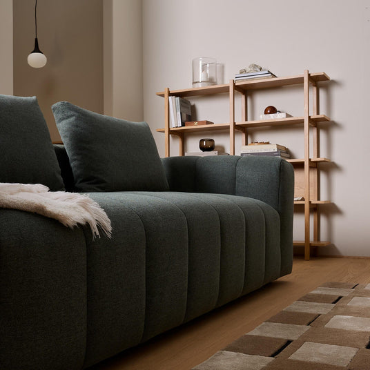 Mörkgrön soffa i närbild med pläd, trähylla med böcker och dekorationer i ett skandinaviskt vardagsrum.