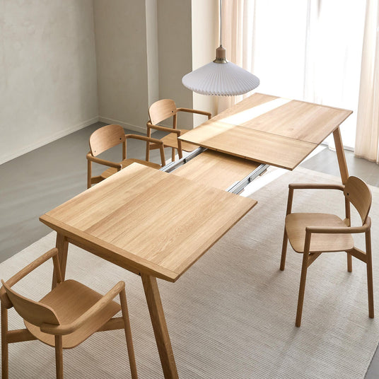 Skandinavisk matplats med ett utdragbart ekbord som är halvvägs utdraget, fyra matchande trästolar, vit plisserad pendellampa, ljus matta och skira gardiner i dagsljus.