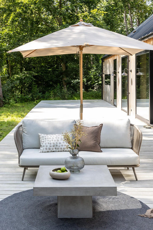 Ljus uteterrass med beige parasoll, soffa med kuddar och betongbord med vas och skål med lime framför ett modernt trähus vid skog.