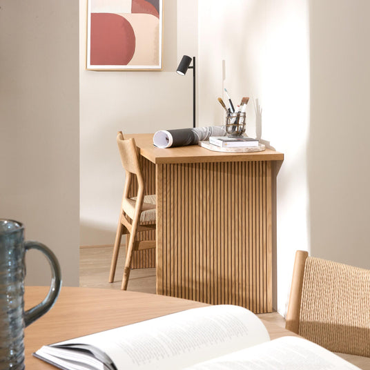 Ljus arbetsplats i trä med ribbad förvaring, stol i naturmaterial, svart bordslampa och konst på väggen; öppna böcker och ritningar på bordet i ett luftigt hem.