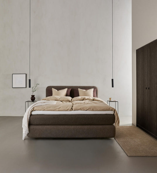 Minimalistiskt sovrum med stoppad säng i bruna toner, beige sängkläder, två svarta pendellampor, små sängbord, väggkonst, vas med gren och garderob i mörkt trä.