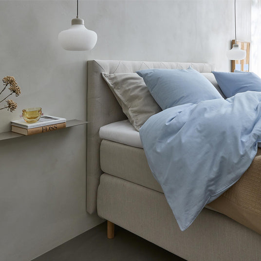 Ljus sovrumsinteriör med beige säng, ljusblått påslakan och kuddar, två vita pendlar samt en smal hylla med vas, gul kopp och boken FOSS bredvid sängen.