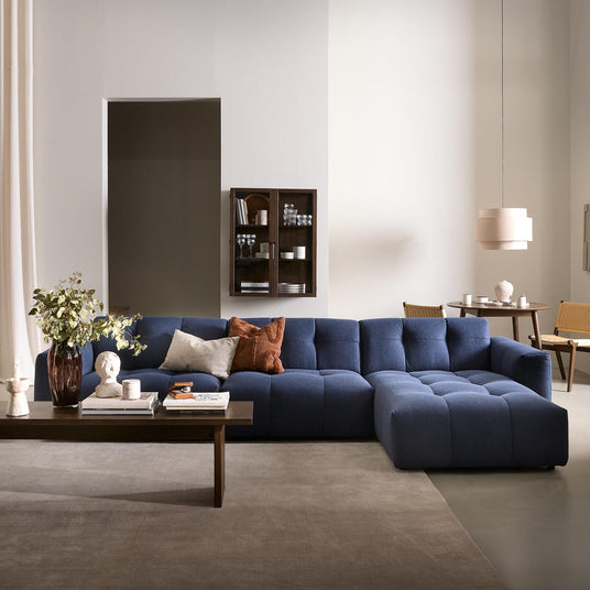 Stilren vardagsrumsmiljö med blå modulsoffa, mörkt träbord med vas och böcker, beige gardiner och en pendellampa i en ljus, neutral palett.