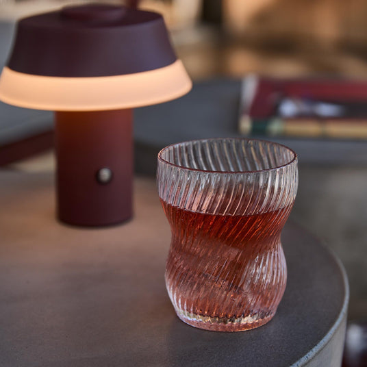 Räfflat dricksglas med röd dryck på ett runt bord bredvid en liten vinröd portabel lampa med varmt sken.