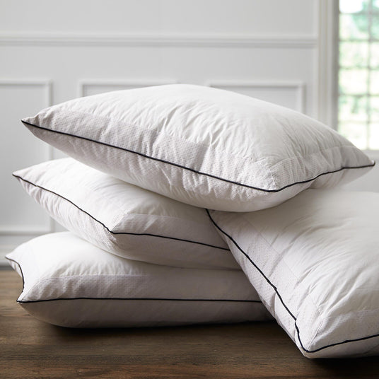 Stapel med fyra vita kuddar med svart passpoal på ett trägolv i ett ljust rum.