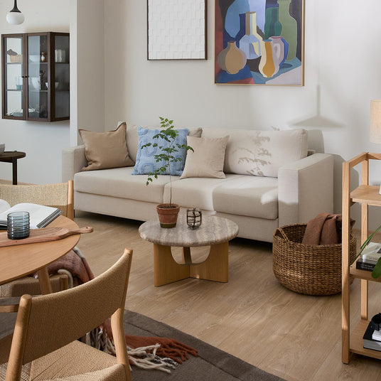 Ljus skandinavisk vardagsrumsmiljö med beige soffa, träbord, väggkonst, korg och gröna växter i dagsljus.