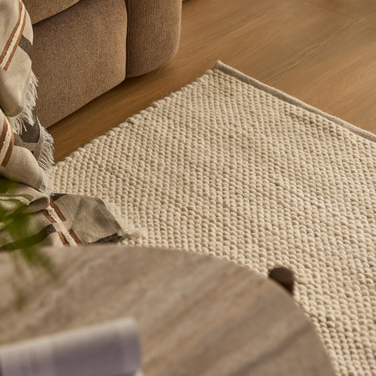 Närbild på ljus, texturerad matta i beige bredvid soffa, pläd och rundt träbord i en varm hemmiljö.