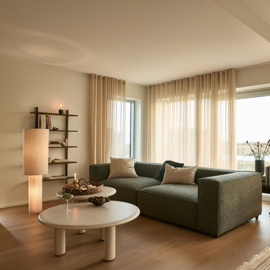Ljus, skandinavisk vardagsrum med grå modulsoffa, runda ljusa soffbord, skira beige gardiner, tända ljus och golvlampa, hylla med böcker samt konsol med skivspelare och högtalare; matta och växt vid stora fönster i dagsljus.
