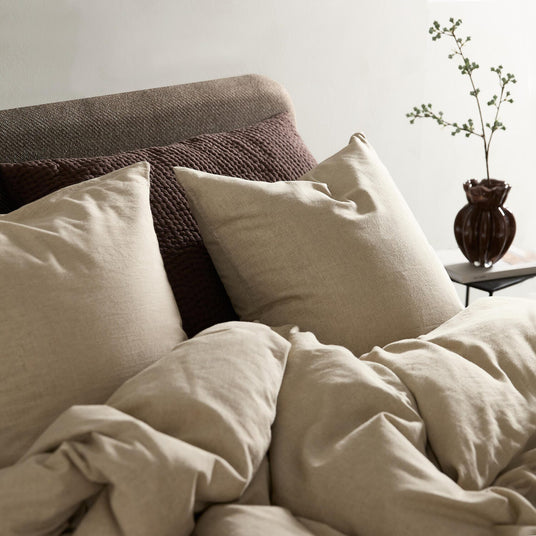 Beige linnebäddset med stora kuddar i en stilren säng; brun texturerad bakgrund och en mörk vas med grön kvist på nattduksbordet.