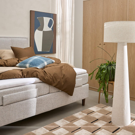 Ljust sovrum med stoppad säng med brunt påslakan, blå rutkudde, textil golvlampa, trägarderob, grön krukväxt och beige geometrisk matta.