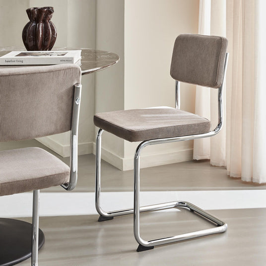 Modern karmfri stol i beige manchester med kromad fribärande ram, intill runt marmorbord med en mörk vas och böckerna ”barefoot living” och ”HOTEL Weekend”.