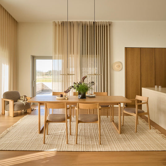 Ljus och luftig matsal i skandinavisk stil med ekbord och fyra stolar på ljus vävd matta, två pendellampor ovanför, skira beige gardiner och en vas med grönt som centerpiece; loungefåtölj vid sidan.