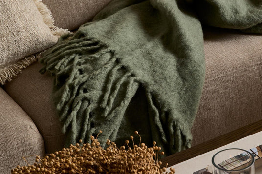 Närbild av en mjuk grön pläd med fransar draperad över en beige soffa med en strukturerad kudde; på soffbordet syns ett ljus i glas och en tidning.