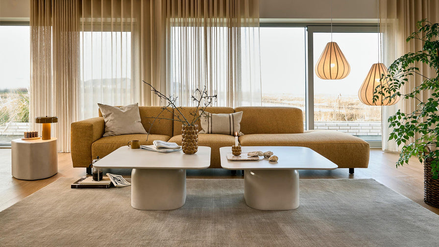 Vardagsrum med senapsgul soffa, skira beige gardiner, två vita soffbord och hängande träpendlar över en stor beige matta, med grön växt vid stora fönster i dagsljus.