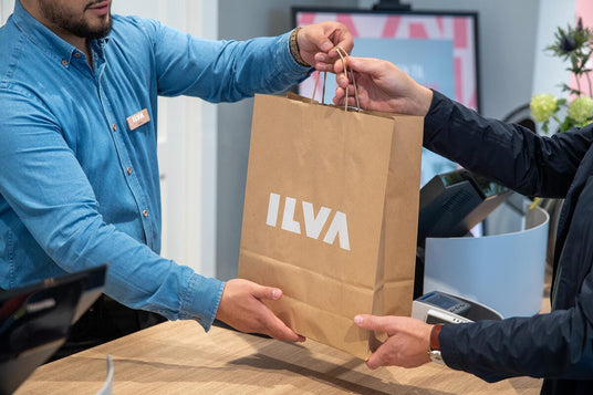 Kund och butikspersonal överlämnar en brun papperspåse med ILVA‑logga vid kassan.