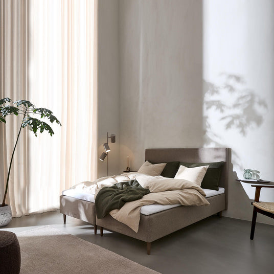 Ljust sovrum med stoppad säng i beige och gröna accenter, mjuka gardiner, vägglampa, sidobord med glasvas och en stor växt i hörnet.