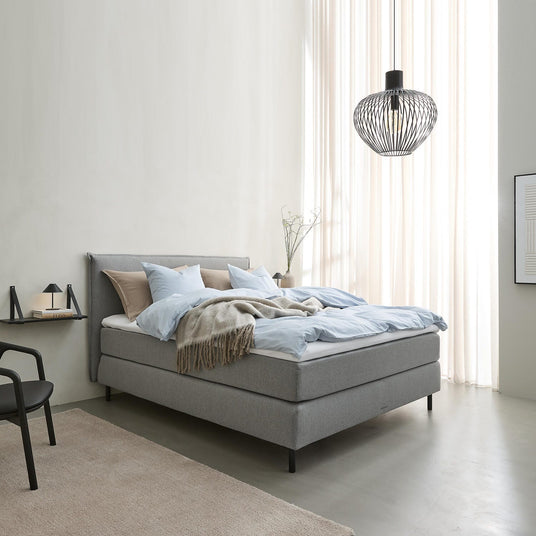 Ljust sovrum i skandinavisk stil med grå klädd säng, ljusblått bäddset och beige pläd, svart trådpendel och tunna gardiner som släpper in dagsljus.