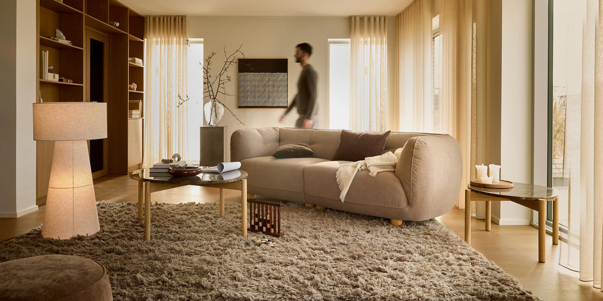 Lugn beige vardagsrum med rundad soffa, mjuk ryamatta, runda soffbord, golvlampa och skira gardiner; en person går förbi.