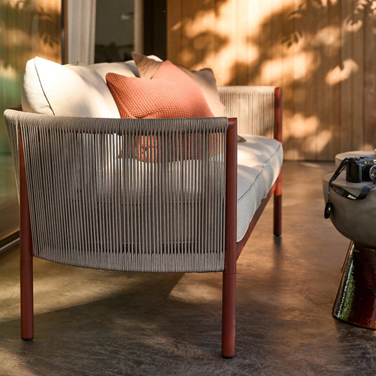 Modern utomhussoffa med grått rep-ryggstöd, roströda ben och beige dynor med terrakottafärgad kudde på terrass i solljus; kamera på sidobord.