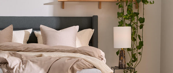 Mysig sovrumsdel med grå stoppad säng, beige bäddning och pläd, sengebord med lampa och böcker, hylla med dekor och hängande växt samt grön sittpuff.