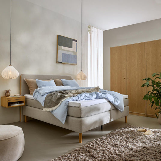 Lugn skandinavisk sovrumsmiljö med ljusgrå säng, ljusblått bäddset och beige pläd, två hängande lampor, svävande sängbord, väggkonst, garderob i trä, ryamatta och grön växt i mjukt dagsljus.