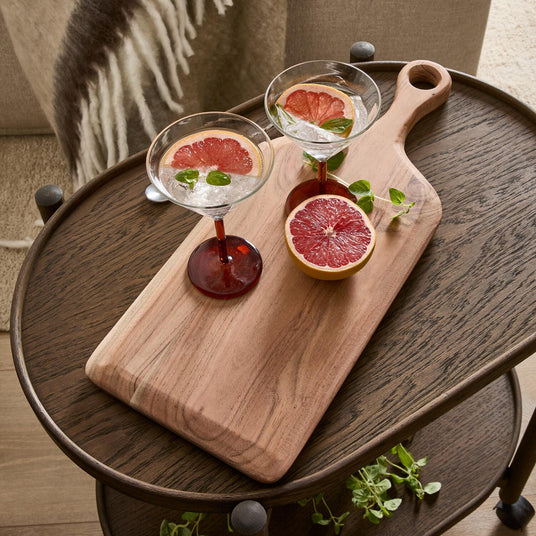 Två cocktailglas med grapefruktskivor och örter på en träskärbräda på ett mörkt sidobord, med en halverad grapefrukt bredvid.