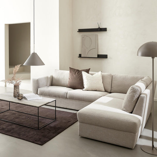 Ljust minimalistiskt vardagsrum med beige hörnsoffa med schäslong, brun matta, rektangulärt soffbord i sten och svart metall, svart läderfåtölj med fotpall, pendel- och golvlampa samt vägghyllor med abstrakt konst.