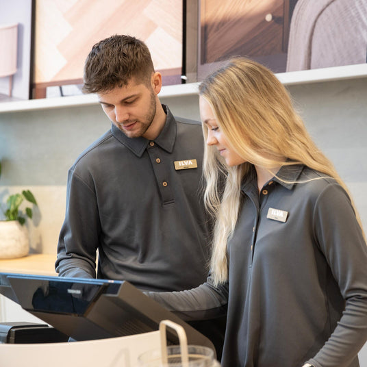 Två butikssäljare i grå ILVA-uniform vid kassadisk som tittar på en kassaterminal i modern showroom-miljö med gröna växter i bakgrunden.