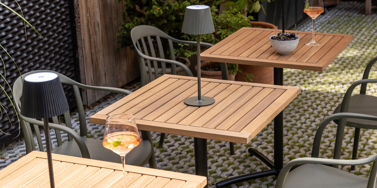 Uteplats med kvadratiska träbord, portabla bordslampor och gröna stolar; glas med rosé och en skål med druvor i dagsljus.