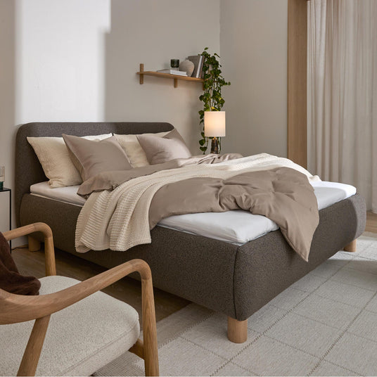 Sovrum med stoppad grå säng, beige bäddset och stickad pläd, ljusa gardiner och vägghylla med växt och lampa i mjukt dagsljus.