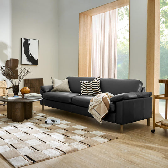 Modern skandinaviskt vardagsrum med svart soffa, geometriskt rutmönstrad matta, kuddar och pläd, trämöbler och stort fönster med ljusa gardiner.