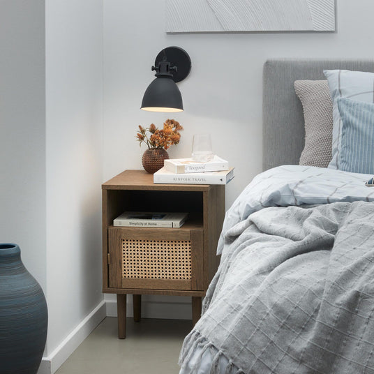 Sovrumshörna med grå säng, blå- och grått bäddset och pläd, ett nattygsbord i trä med rottinglucka, liten vas med torkade blommor, svart vägglampa och en stapel böcker – Kinfolk Travel, Simplicity at Home och Be Toogood – samt ett vattenglas ovanpå.