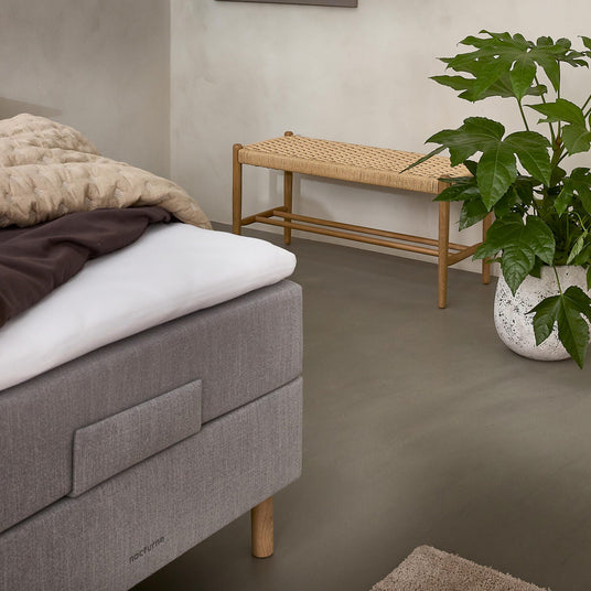 Lugn sovrumshörna med grå stoppad säng med “nocturne”-märke, beige pläd, flätad träbänk och grön krukväxt i vit kruka på neutralt golv.