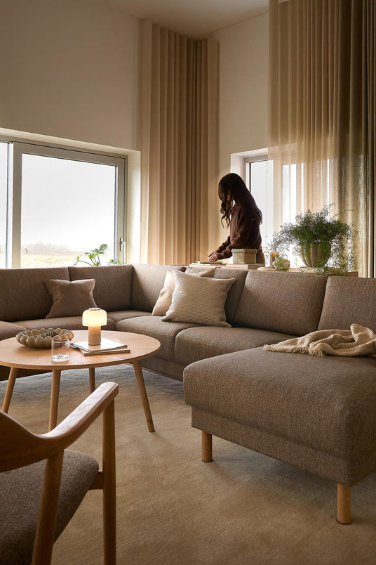 Ljust vardagsrum med beige gardiner, brun tygsoffa i L‑form, runt träbord med liten lampa och filt på schäslongen; en person står vid fönstret.