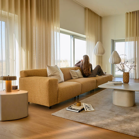 Ljust vardagsrum med skira beige gardiner, senapsgul soffa med kuddar, två vita pendellampor och lågt bord med vas och kvistar på stor matta; en person sitter vid fönstret och blickar ut.