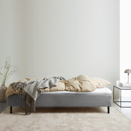 Grå stoppad säng med beige bäddset och grå pläd; nattduksbord med lampa och böcker i ett ljust, minimalistiskt sovrum.