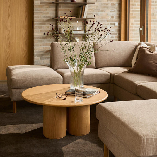 Beige hörnsoffa med runt soffbord i trä, vas med blommor, böcker och ett glas i ett ljust vardagsrum.